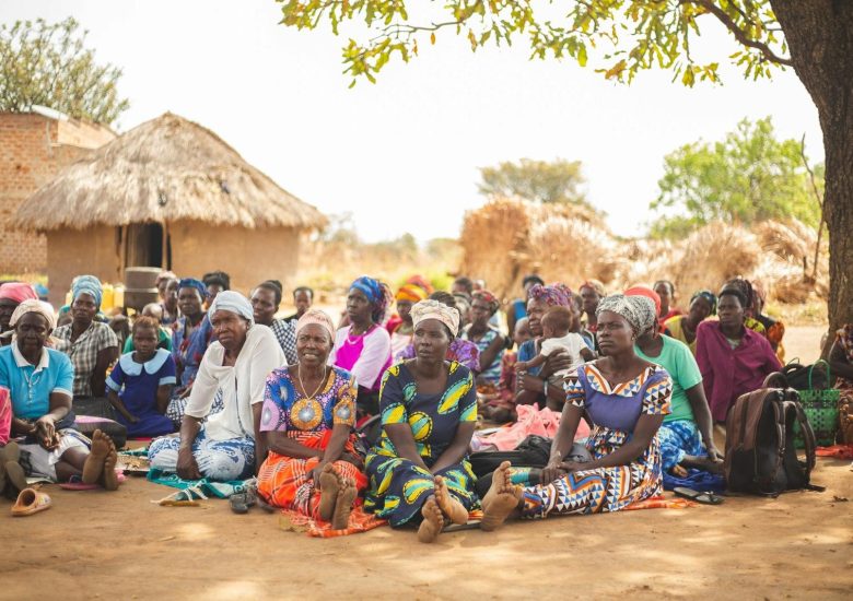 A diverse group gathered under a tree in an African village, showcasing vibrant cultural attire.